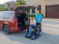 A person stands beside a parked red minivan, operating a lift to load a blue mobility scooter into the vehicle's open trunk. The scene is set in a residential driveway in front of a large house.
