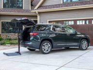A dark green SUV is parked in a driveway with its trunk open, next to a portable lift. The house in the background has a double garage and a beige and brown exterior.