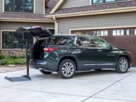 A dark green SUV is parked in a driveway with its trunk open, next to a portable lift. The house in the background has a double garage and a beige and brown exterior.