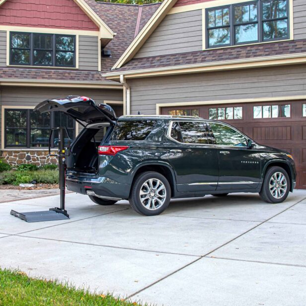 A dark green SUV is parked in a driveway with its trunk open, next to a portable lift. The house in the background has a double garage and a beige and brown exterior.