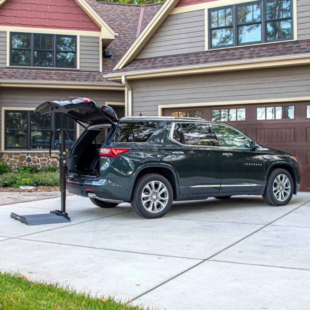 A dark green SUV is parked in a driveway with its trunk open, next to a portable lift. The house in the background has a double garage and a beige and brown exterior.