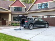 A black SUV is parked in the driveway of a suburban house with an open hatch and a hitch-mounted lift, extending behind the vehicle. The house has a stone and wood exterior with a two-car garage.