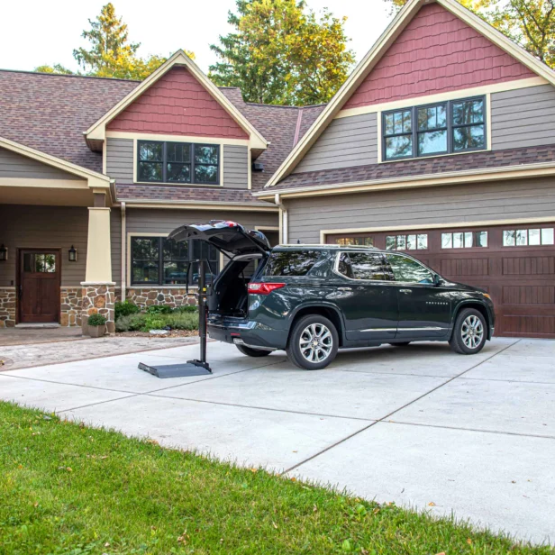 A black SUV is parked in the driveway of a suburban house with an open hatch and a hitch-mounted lift, extending behind the vehicle. The house has a stone and wood exterior with a two-car garage.