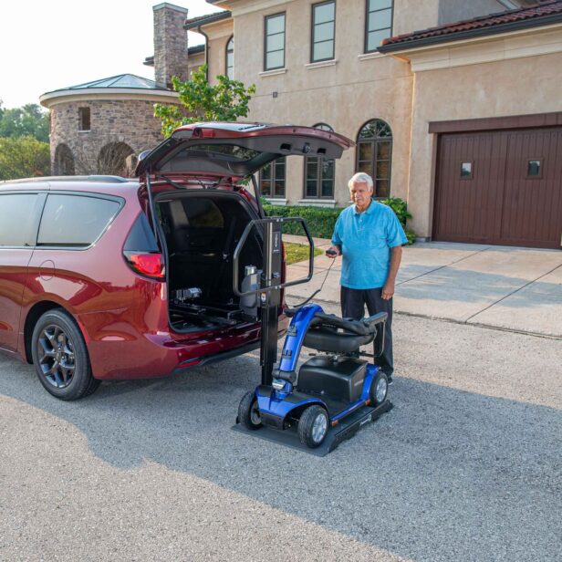 A person stands beside a parked red minivan, operating a lift to load a blue mobility scooter into the vehicle's open trunk. The scene is set in a residential driveway in front of a large house.