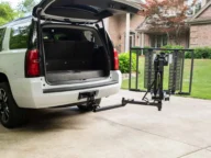 A white SUV with an open trunk is parked in a driveway. A foldable platform lift is attached to the rear hitch of the SUV, extending out towards the ground. A brick house is in the background.
