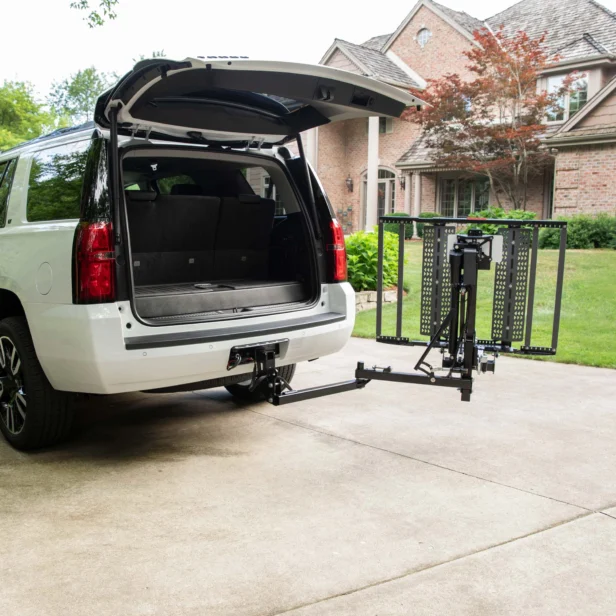 A white SUV with an open trunk is parked in a driveway. A foldable platform lift is attached to the rear hitch of the SUV, extending out towards the ground. A brick house is in the background.