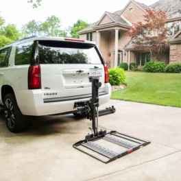 A white SUV with a rear-mounted lift platform is parked in a driveway in front of a suburban house. The lift is folded and attached to the vehicle's tow hitch.