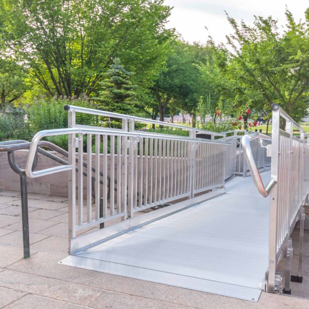 Metal ramp with railings provides wheelchair access to a raised area in a park, surrounded by greenery and trees.