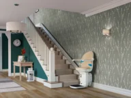 A stairlift is installed along a carpeted staircase in a modern home interior with patterned wallpaper, a clock, a small table, and a light fixture.