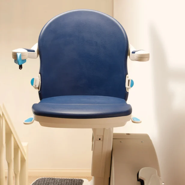A blue stairlift installed on a staircase, featuring a padded seat, armrests, and a control lever on the armrest.