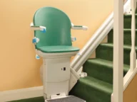 A green stairlift installed along a carpeted staircase, featuring a cushioned seat, armrests, and a footrest for safe and easy accessibility.