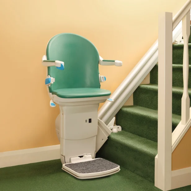 A green stairlift installed along a carpeted staircase, featuring a cushioned seat, armrests, and a footrest for safe and easy accessibility.