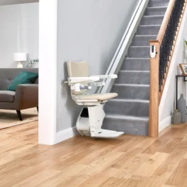 A beige stairlift is installed on a carpeted staircase in a modern living room with a gray sofa, teal pillows, a wooden floor, and a small side table with decor.