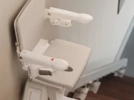 A beige stairlift is attached to a staircase, featuring a padded seat, armrests, and a safety belt.
