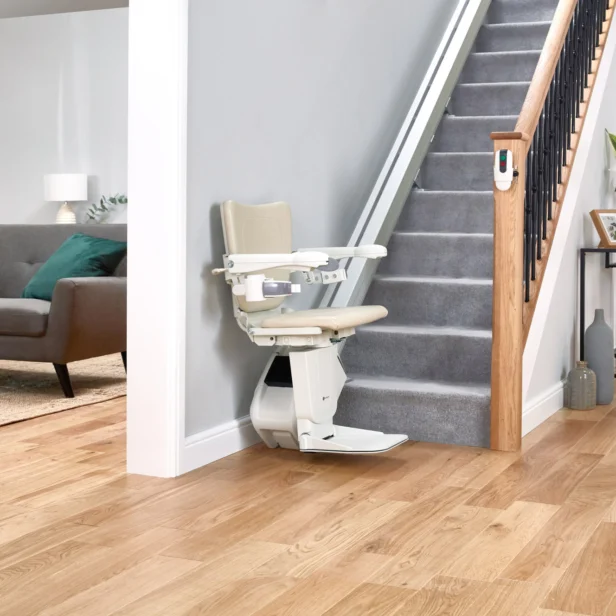 A beige stairlift is installed on a carpeted staircase in a modern living room with a gray sofa, teal pillows, a wooden floor, and a small side table with decor.