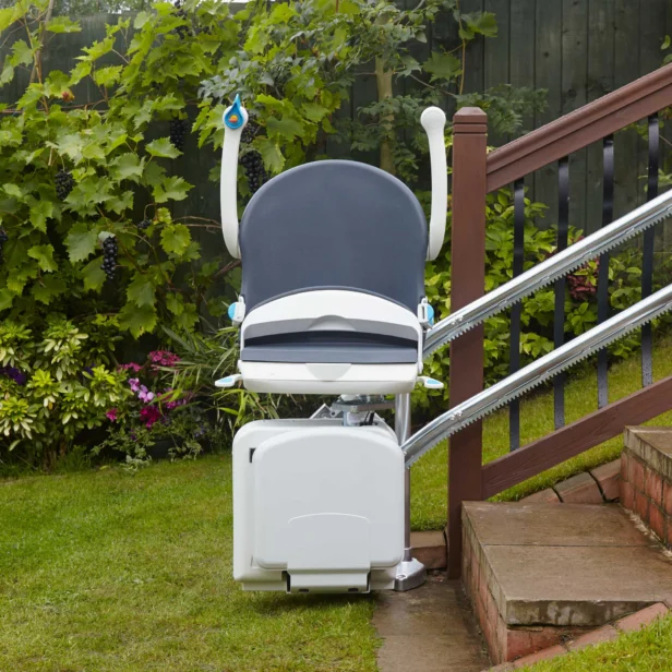 Outdoor stair lift installed beside a wooden staircase in a garden, with greenery and flowers in the background.