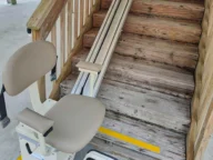 Image of a stair lift installed beside a wooden staircase, intended for assisting individuals with mobility difficulties to ascend and descend the stairs.