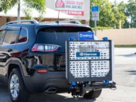 A car is parked with a wheelchair lift attached to its rear. There is a building in the background with a sign indicating curbside pickup is available.