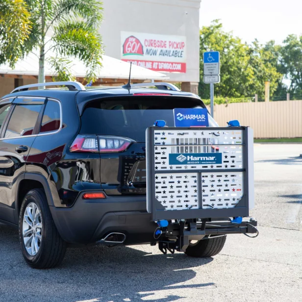 A car is parked with a wheelchair lift attached to its rear. There is a building in the background with a sign indicating curbside pickup is available.