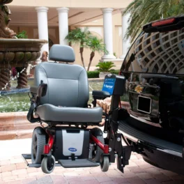 A motorized wheelchair is mounted on a lift attached to the back of a black SUV, parked near a fountain and a building with columns.