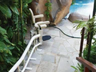 A chair lift is installed on a stone pathway leading to a pool surrounded by rocks and lush greenery.