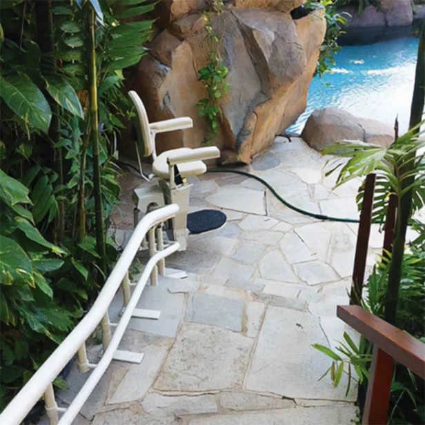 A chair lift is installed on a stone pathway leading to a pool surrounded by rocks and lush greenery.