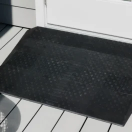 A black rubber doormat with a textured pattern is placed in front of a white door on a light gray wooden deck.