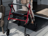 A person using a red walker is entering a house via a ramp from a wooden deck. The walker has a seat and four wheels.