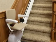 A stair lift is installed on a carpeted staircase with wooden railings, featuring a padded seat, armrests, and a footrest.