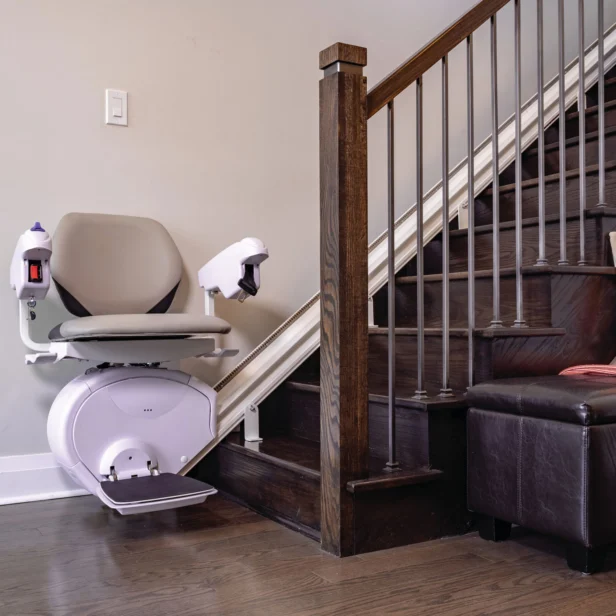 A white and gray stair lift is installed along a wooden staircase next to a gray wall. An armchair is partially visible next to the staircase.