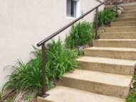 Concrete outdoor steps with a metal railing on the left side, leading up to a beige building with a small window. Green plants are growing alongside the steps.