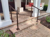 Front porch with brick steps, white columns, and two metal railings. There is a white swing with coral cushions and a small white side table. Bushes and a path lead up to the porch.