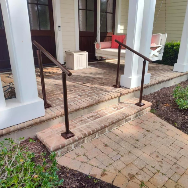Front porch with brick steps, white columns, and two metal railings. There is a white swing with coral cushions and a small white side table. Bushes and a path lead up to the porch.
