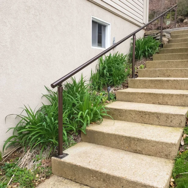 Concrete outdoor steps with a metal railing on the left side, leading up to a beige building with a small window. Green plants are growing alongside the steps.