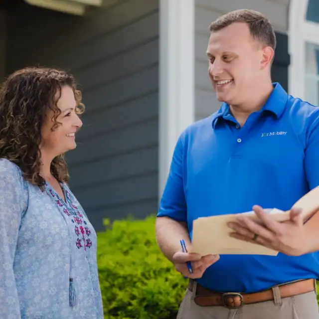 A woman in a blue blouse smiles while talking to a man in a blue polo shirt holding a clipboard and pen outside a house.