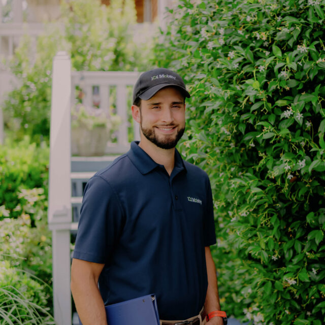 A man in a navy blue uniform and cap stands outdoors near lush greenery, holding a blue clipboard. He has a beard and smiles at the camera.