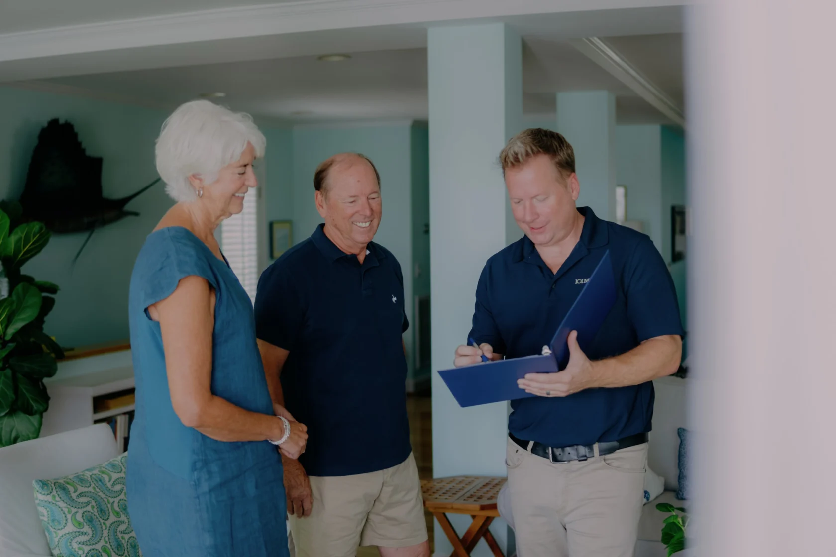 Three adults, two standing and one holding a clipboard, are conversing in a room with light blue walls and decor.