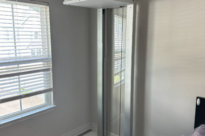 A small residential vertical lift installed in a corner of a room near a window, with a closed hatch on the floor and control panel at the top.