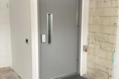 A closed gray metal door with a window in an industrial building, featuring an accessible ramp and partially exposed insulation above the frame.