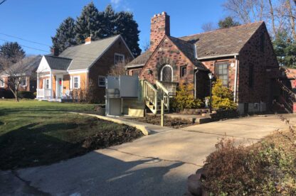 A brick house with a staircase and porch, tall chimney, and a driveway in front. Another house is partially visible on the left. Both houses have yards with patches of grass and some leafless trees.