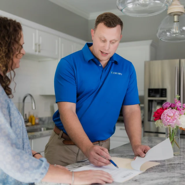 A man in a blue polo and a woman are reviewing documents on a kitchen countertop. The man is holding a pen and pointing at the papers.