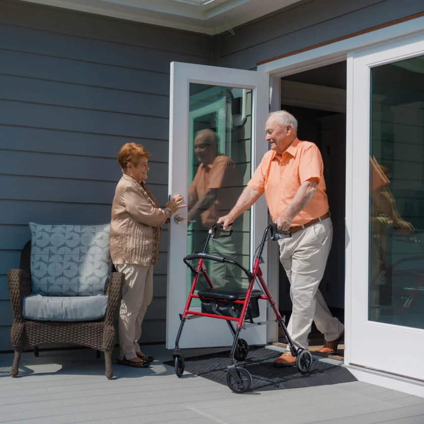 An elderly man using a walker steps out of a house through a sliding glass door while an elderly woman holds the door open for him. A wicker chair with a cushion is visible on the deck.