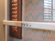 A white strip with four round buttons in different colors (red, green, blue, and white) is mounted on a patterned wallpaper background with wooden window shutters to the left.