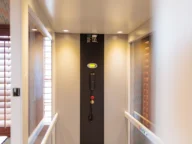 Interior of a residential elevator with a control panel, emergency phone, and handrails. Sunlight filters through wooden shutters on either side.