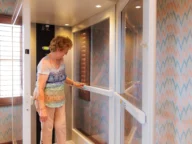An elderly woman operates a home elevator inside a residential setting with patterned wallpaper and a window with blinds.