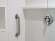 A bathroom shower with white tile walls, a stainless steel grab bar, and a single-lever handle for the shower controls.