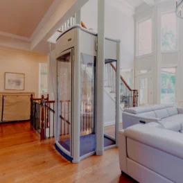 A small glass elevator is situated in the center of a spacious, brightly lit living room with a large gray sectional sofa, wooden floors, and a staircase leading to an upper level.