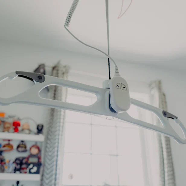 Overhead patient lift hangs from the ceiling by a cord in a room with shelves and a window in the background.