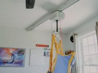 An overhead patient lift with a blue and yellow harness is mounted in a room with a calendar, poster, and window.