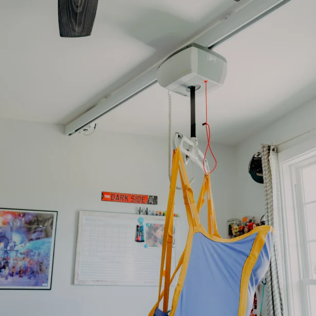 An overhead patient lift with a blue and yellow harness is mounted in a room with a calendar, poster, and window.